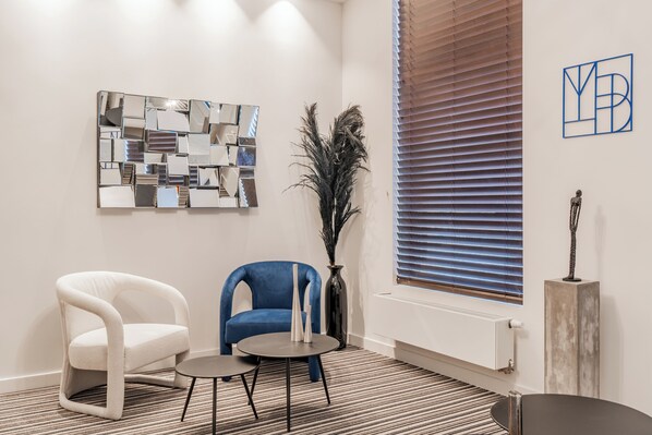 Quarto moderno com cadeira branca e azul, uma mesinha e um espelho decorativo na parede.