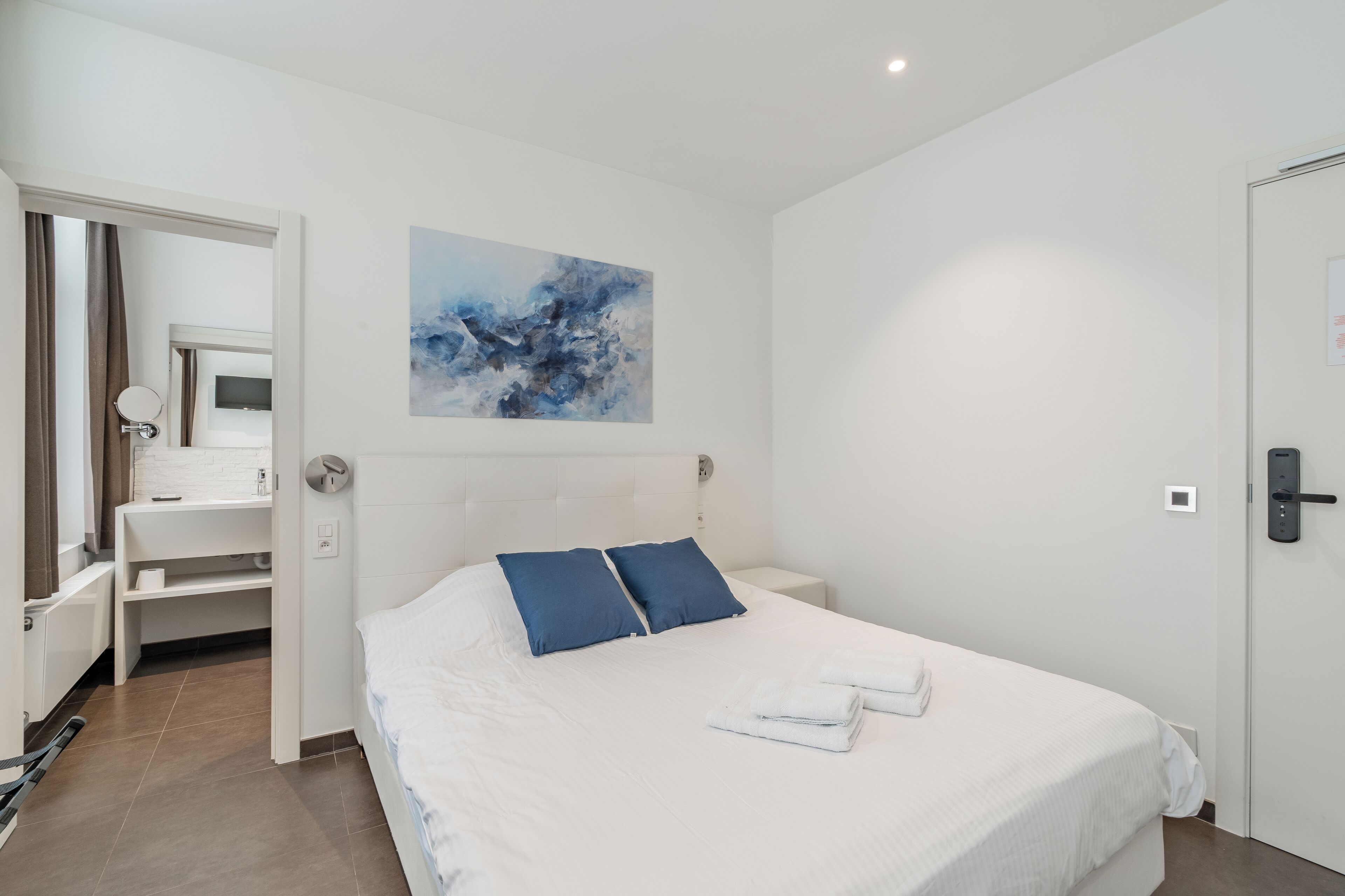 Une chambre d’hôtel avec un grand lit, une tête de lit blanche, un tableau abstrait bleu, une table de chevet blanche et une salle de bain visible à travers une porte ouverte.