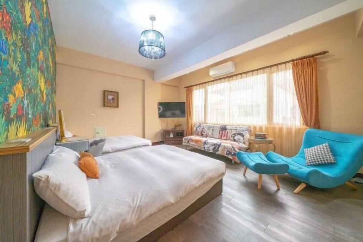 A hotel room with a bed, a blue armchair, a sofa, a television, and a colorful painting on the wall.