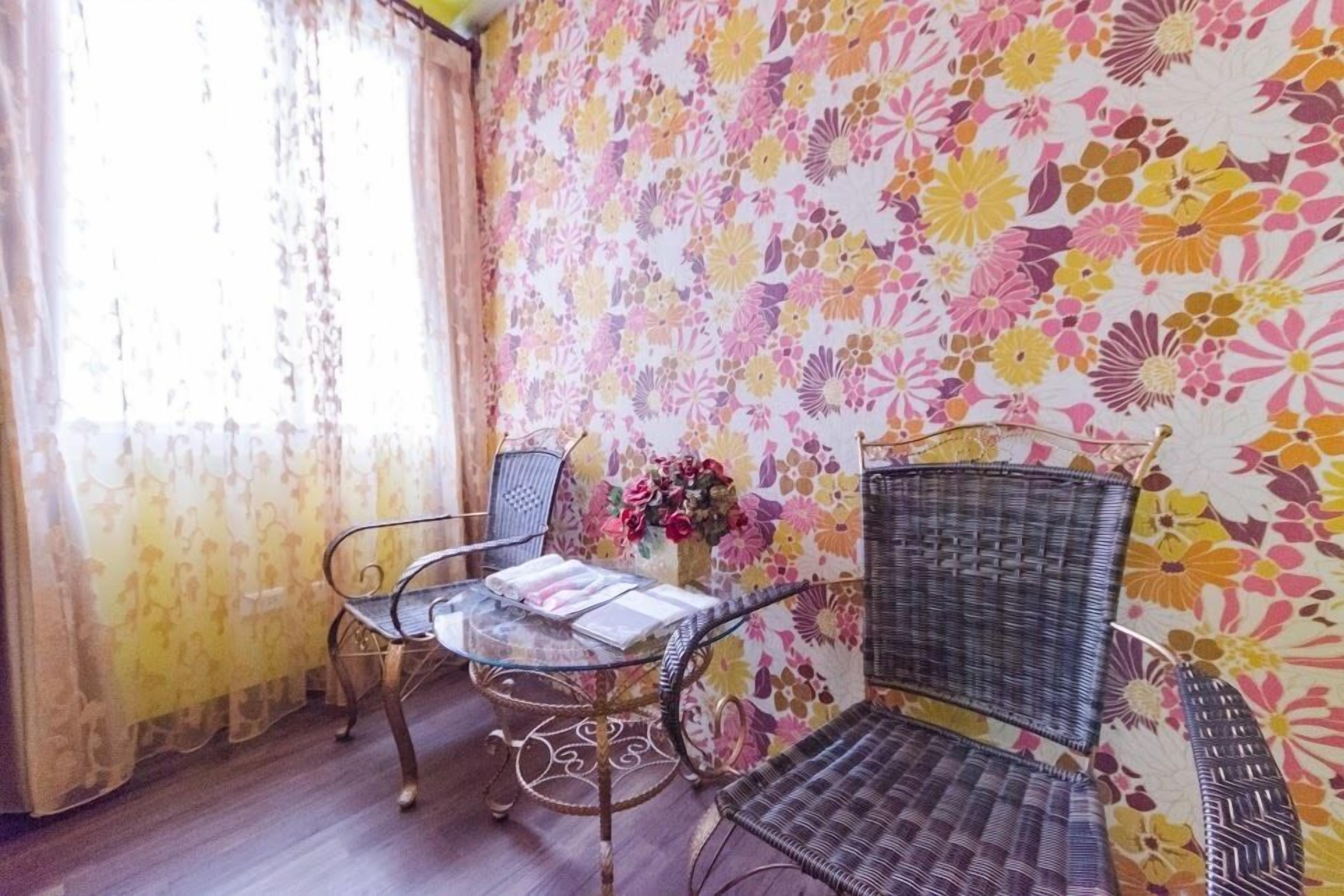 A room with a floral wallpaper, a glass table with chairs, and a window with curtains.