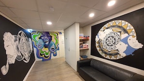 Un intérieur moderne avec un canapé en cuir noir, un plancher en bois et des œuvres d’art murales aux couleurs vives, notamment un motif circulaire et une fresque représentant des visages abstraits.