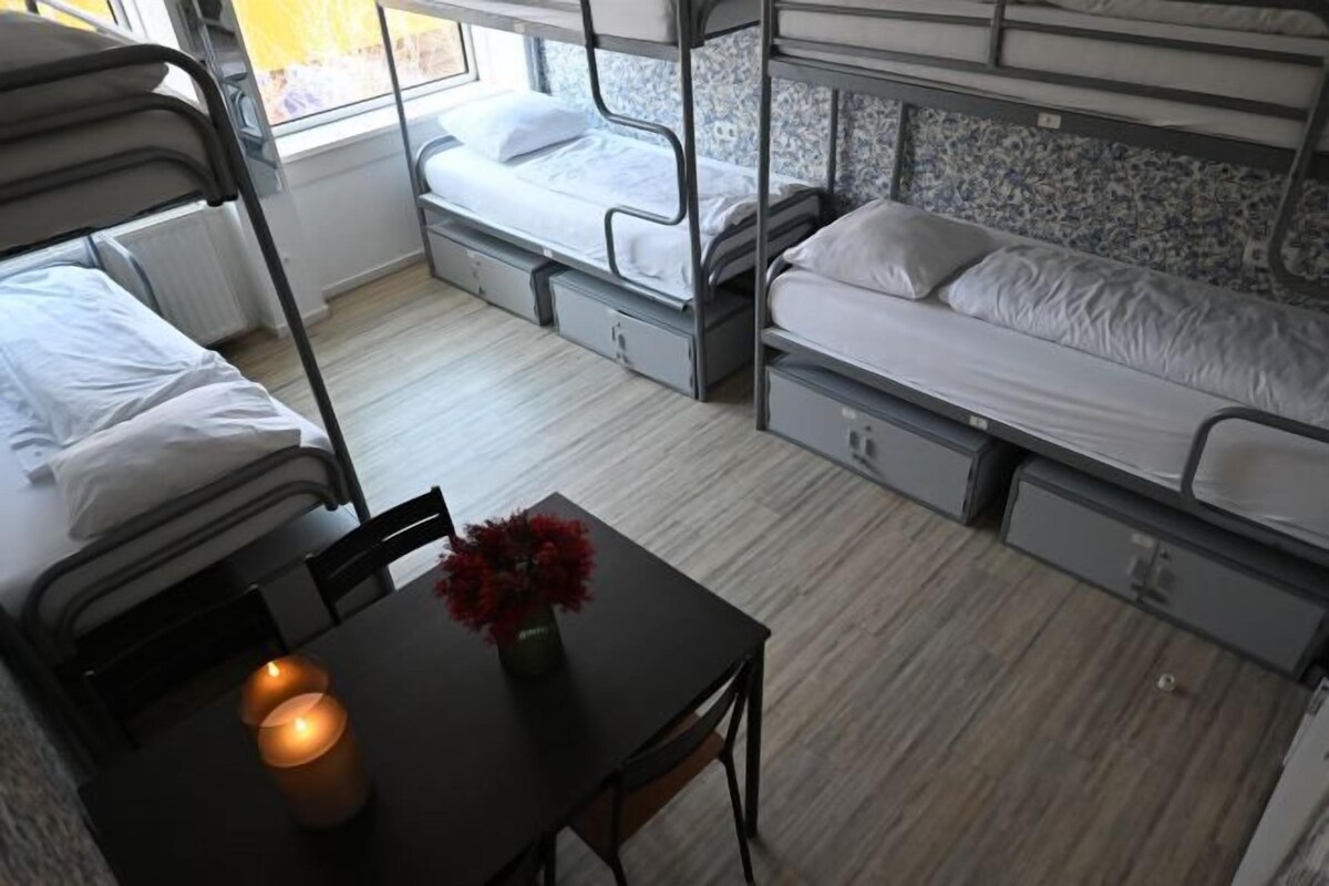 A dormitory room with bunk beds, a small table with a candle, and a window with a view.