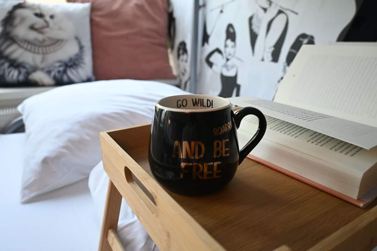 Una bandeja de madera con una taza negra y un libro abierto sobre una cama.
