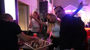 Un groupe de personnes joue aux échecs dans une pièce faiblement éclairée, autour d’une table en bois sur laquelle se trouvent une bouteille de vin et un gobelet rouge.