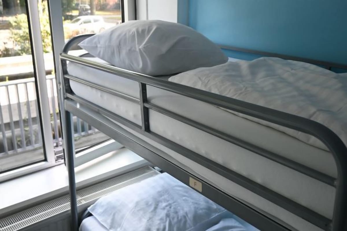A metal bunk bed with white bedding, a window with a view of a street, and a blue wall.