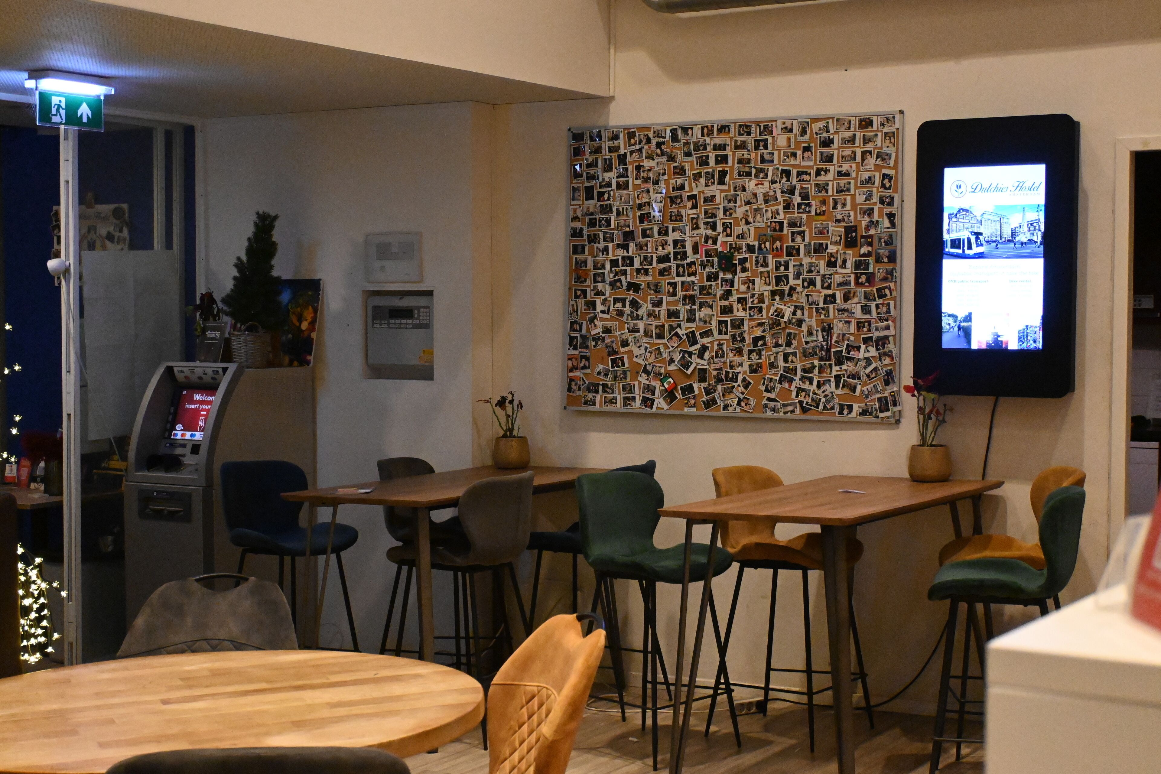A hotel lobby with a wall decorated with photos, a digital screen displaying hotel information, and several wooden tables and chairs.