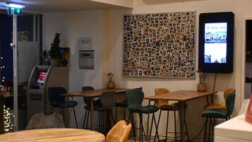 A hotel lobby with a wall decorated with photos, a digital screen displaying hotel information, and several wooden tables and chairs.