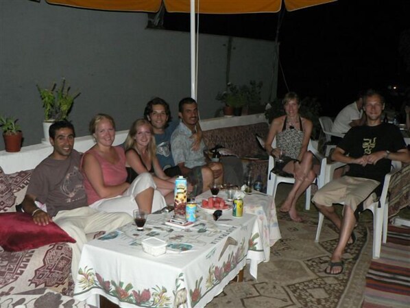 A group of people seated around a table with a white tablecloth, surrounded by potted plants and a yellow umbrella.