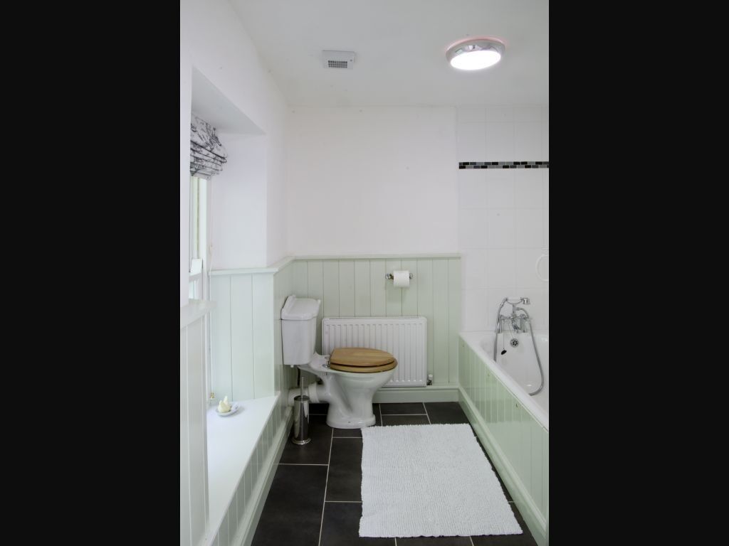 Casa de banho com sanita, radiador branco, banheira e tapete branco no chão.