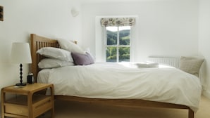 A bedroom with a wooden bed, white bedding, a nightstand with a lamp, and a window with a view of greenery. - Bronfelin & Troed-y-rhiw Holiday Cottages (Llanwrtyd Wells)