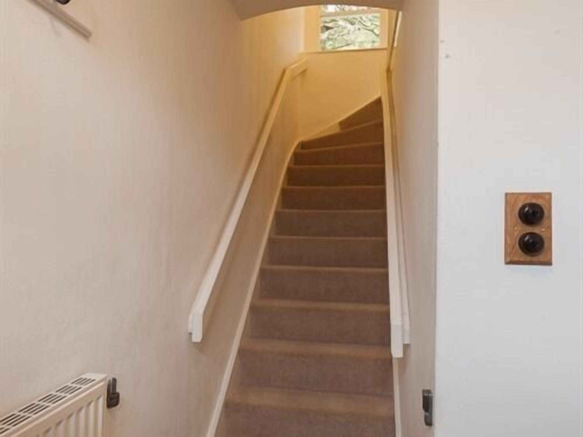 A staircase with carpeted steps and a handrail, leading to a landing with a light switch and a vent.