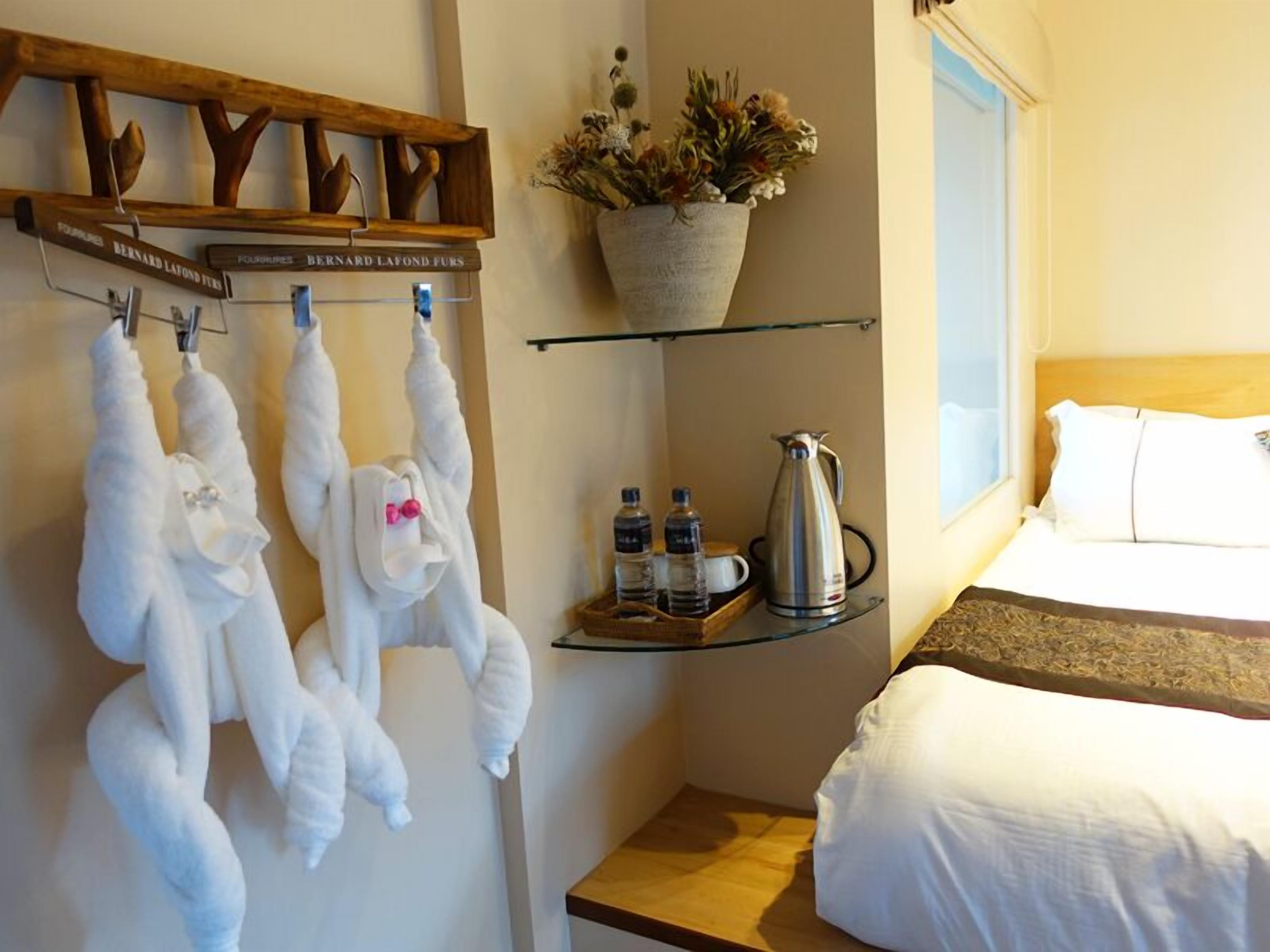 A hotel room with a bed, a shelf with a coffee pot and water bottles, a wall-mounted towel rack with plush toys, and a potted plant.