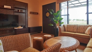 Una sala de estar con televisión, muebles de mimbre, un ventanal grande y una planta.