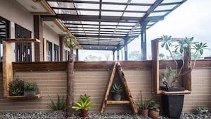Un espacio exterior techado con un banco de madera, plantas en macetas y una celosía metálica.