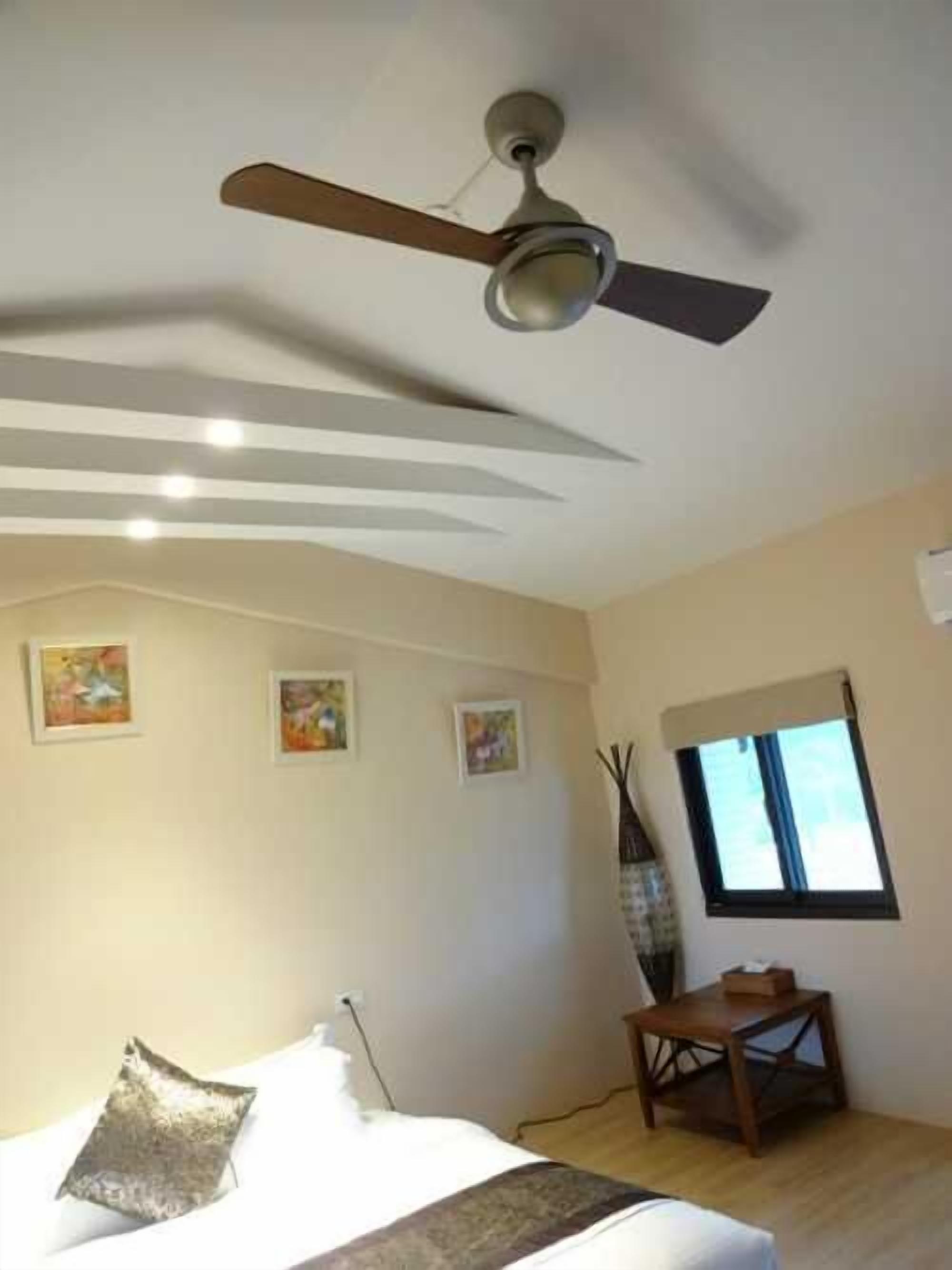 Une chambre à coucher avec un lit, un ventilateur de plafond, des œuvres d’art accrochées au mur et une fenêtre.