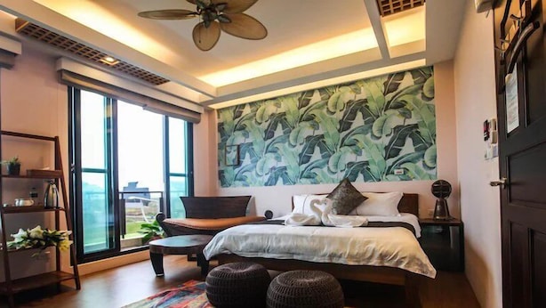 Un dormitorio moderno con un ventanal grande, un ventilador de techo, una cama con un mural de hoja de plátano y una mesita de noche de madera.