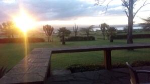 A patio with a stone table and a view of a grassy field with trees and a sunset in the background. - Jacaranda Lake Elementaita Lodge (Gilgil)