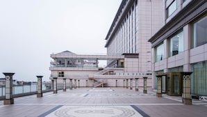 Un espacio exterior amplio con piso estampado, una escalera que conduce a un nivel superior y un edificio moderno con grandes ventanales.