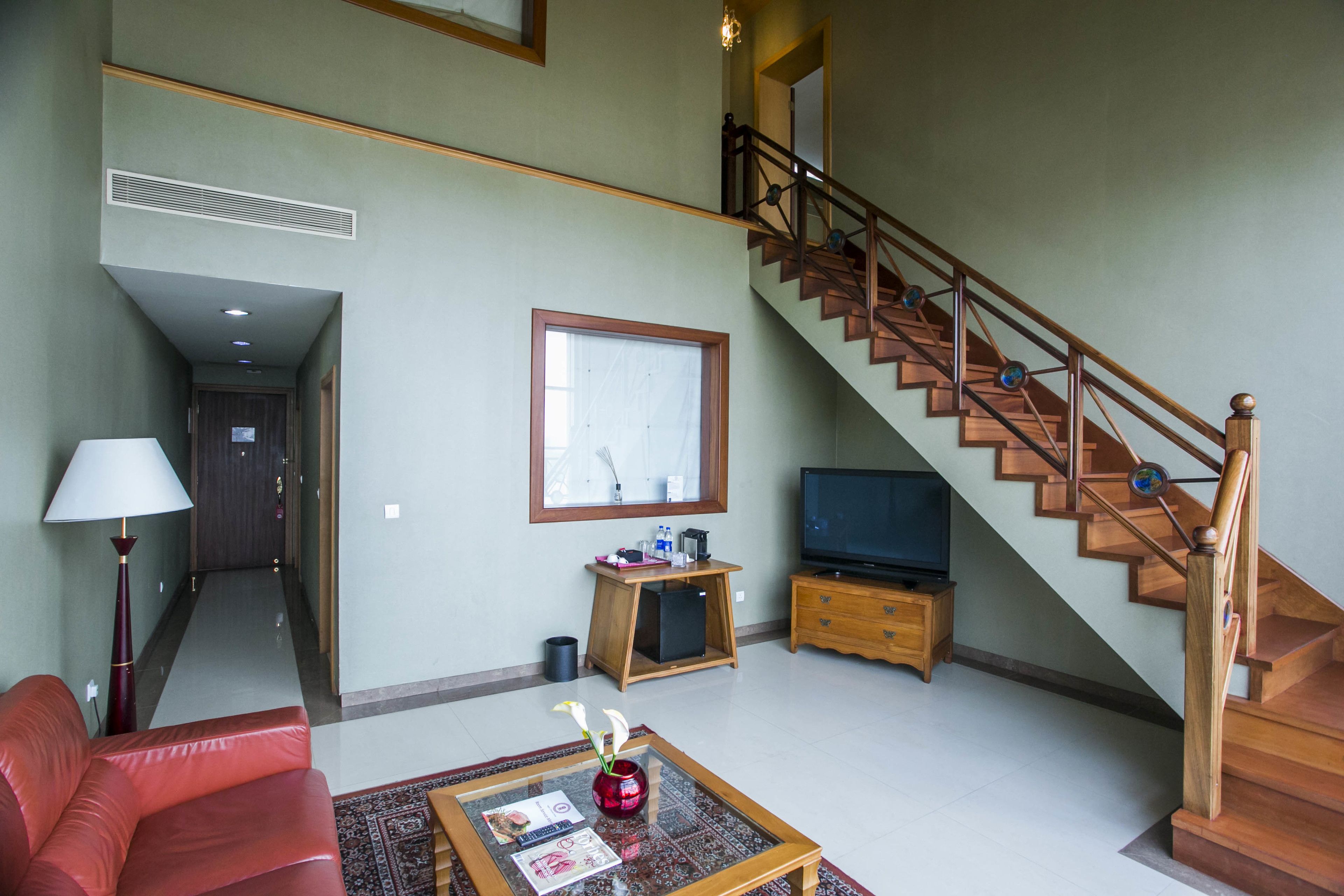Un salón amplio con un sofá rojo, una mesa de centro de vidrio con revistas, un mueble de televisión de madera y una escalera con barandilla de madera.