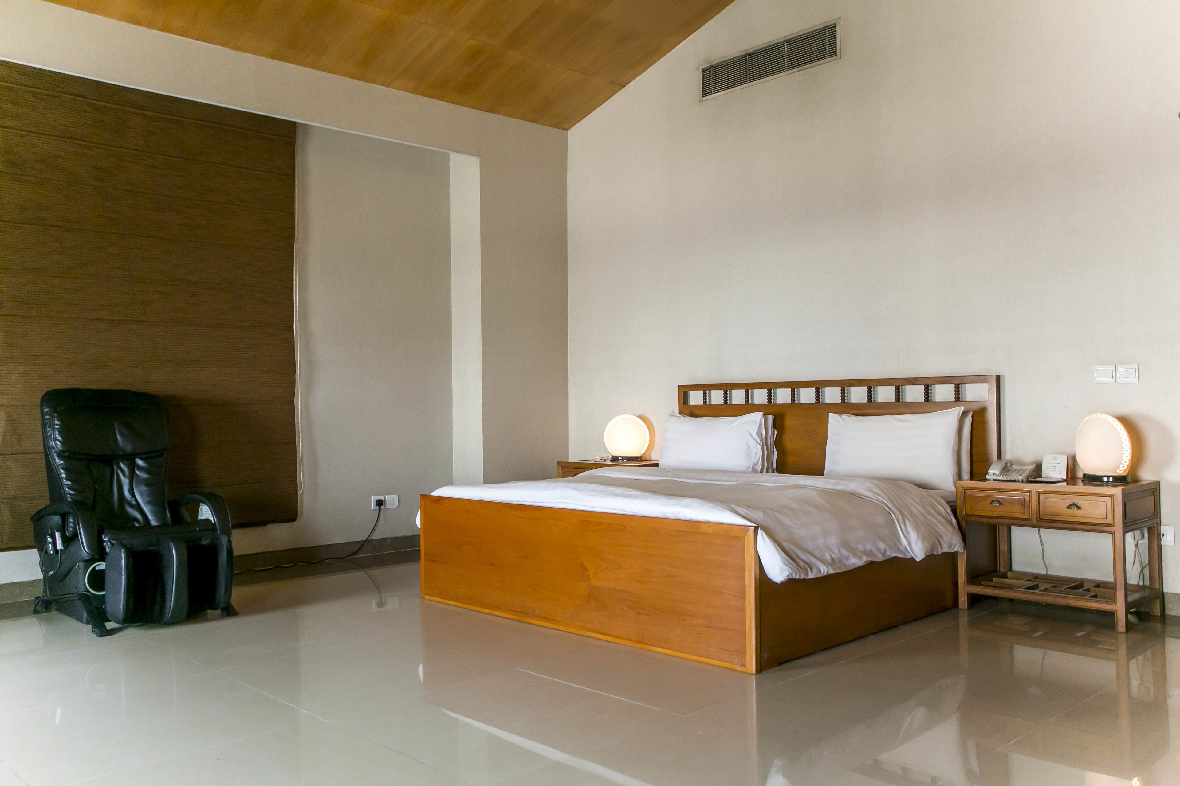 A modern bedroom with a wooden bed, bedside lamps, a massage chair, and a wooden nightstand.