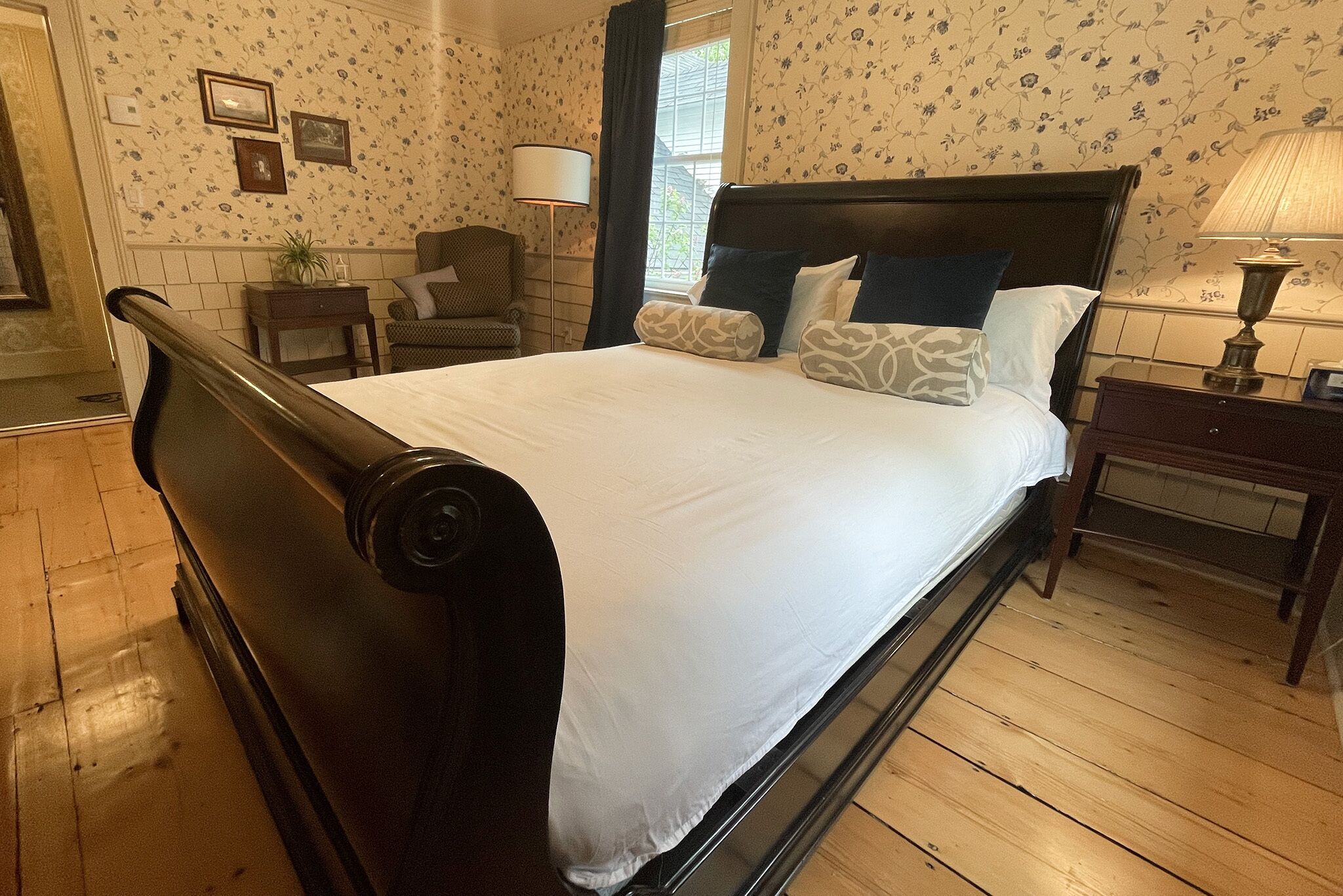 Una cama bien tendida con un cabecero oscuro, una mesita de noche con lámpara y una silla en una habitación con papel tapiz floral y suelo de madera.