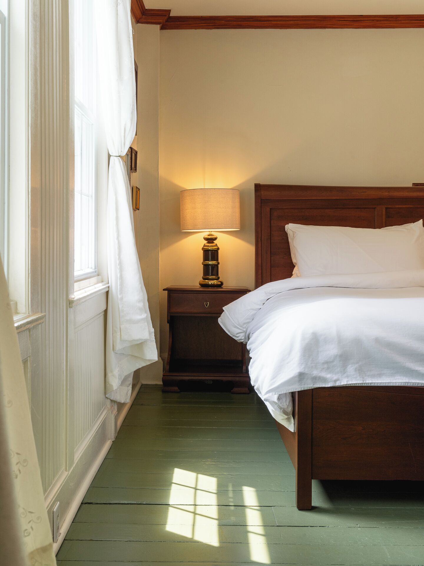 A bedroom with a wooden bed, a nightstand with a lamp, a window with curtains, and a green wooden floor.