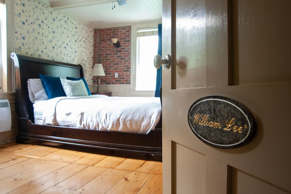 A bedroom with a wooden floor, a bed with white bedding and blue pillows, a brick wall, and a door with a nameplate reading "William Lee".