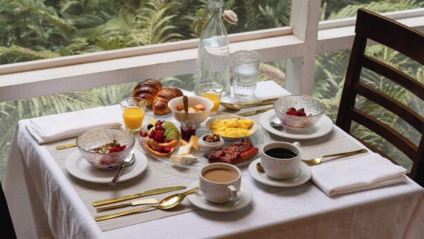 Una mesa de desayuno preparada con croissants, fruta, huevos revueltos y café, con vistas al verdor exterior.