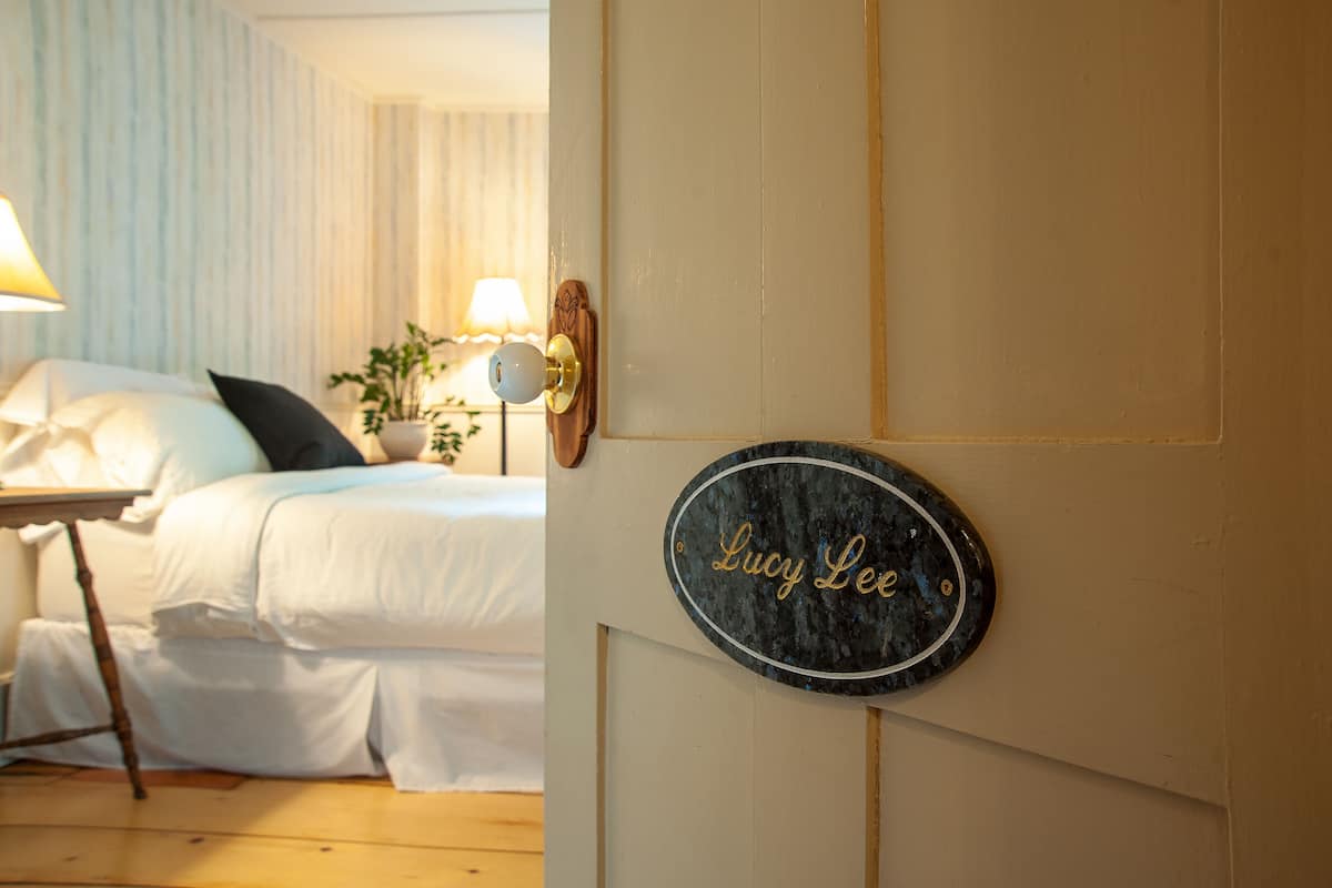 A neatly made bed with white linens and a small bedside table with a lamp. A sign with the name "Lucy Lee" is mounted on the wall.