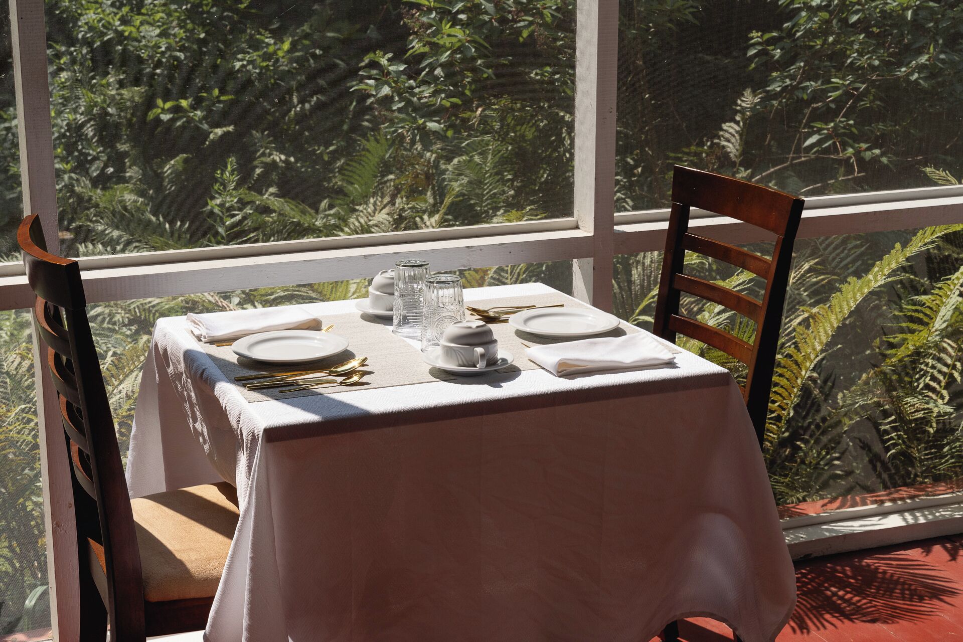 A dining table set for two with white tablecloth, plates, cutlery, and glasses, positioned by a window with a view of tropical foliage.