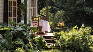 Una casa de dos pisos con un balcón verde, un área de comedor con mesa y sillas, y abundante vegetación alrededor de la propiedad.