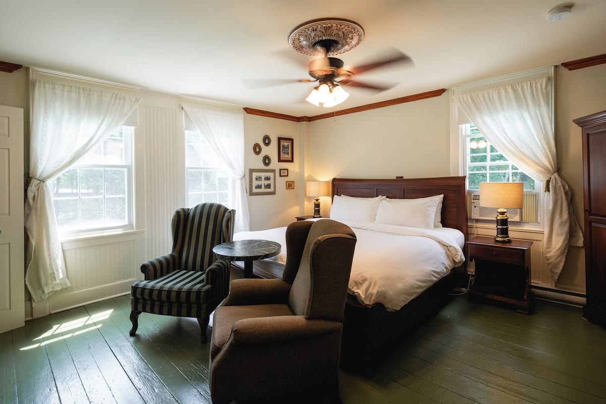 A hotel room with a bed, two armchairs, a small table, a ceiling fan, and a window with curtains.