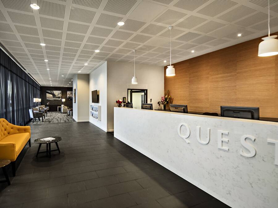 Un área de recepción de hotel moderna con un mostrador de mármol con el rótulo “QUEST”, un sofá amarillo y una pared de acento de madera.