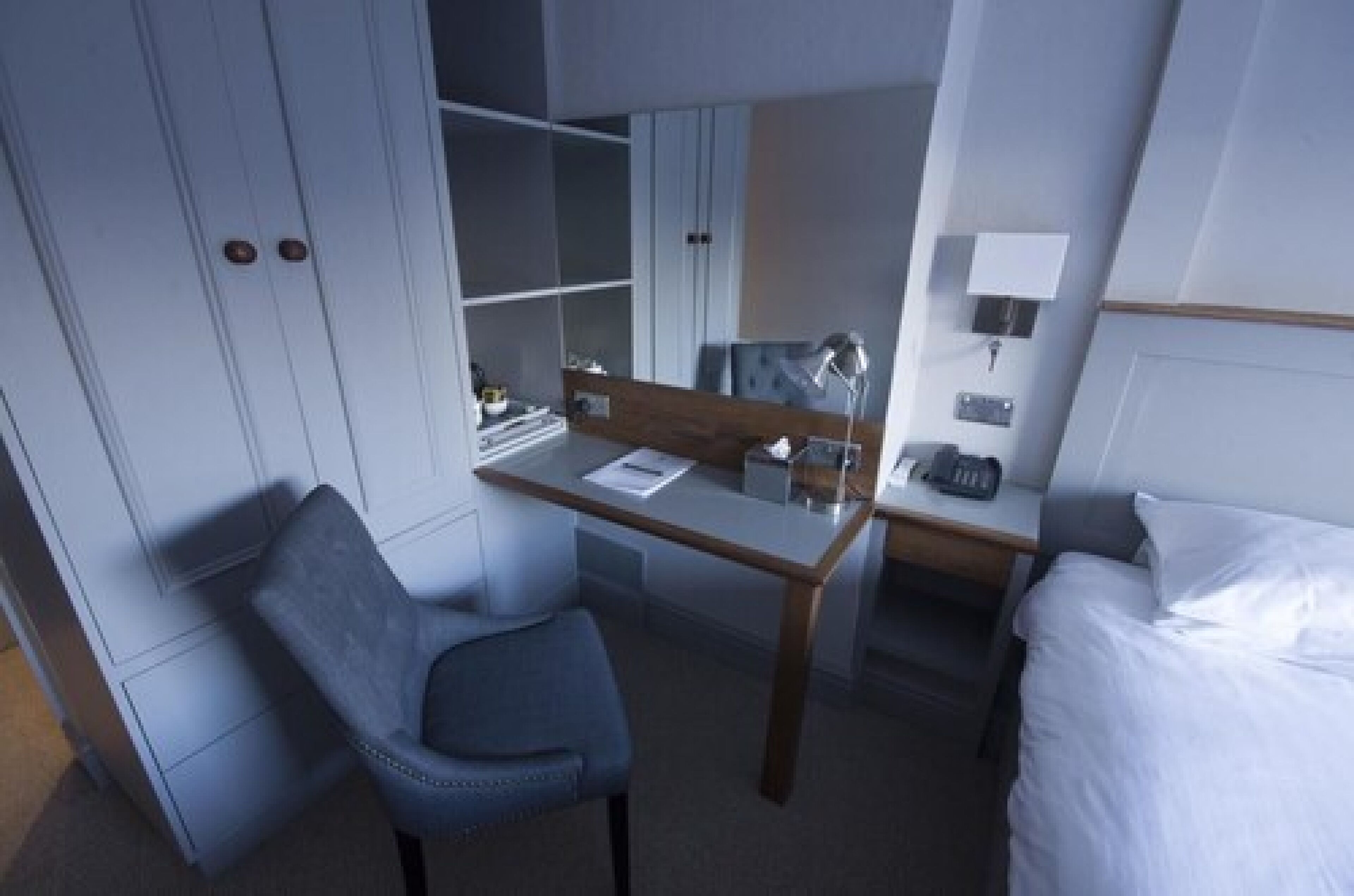 A hotel room with a bed, a desk, a chair, a wardrobe, and a bedside table with a lamp.