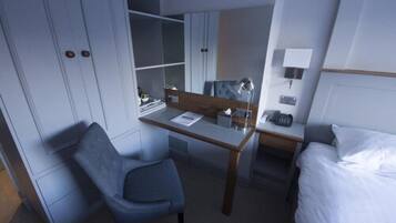A hotel room with a bed, a desk, a chair, a wardrobe, and a bedside table with a lamp.