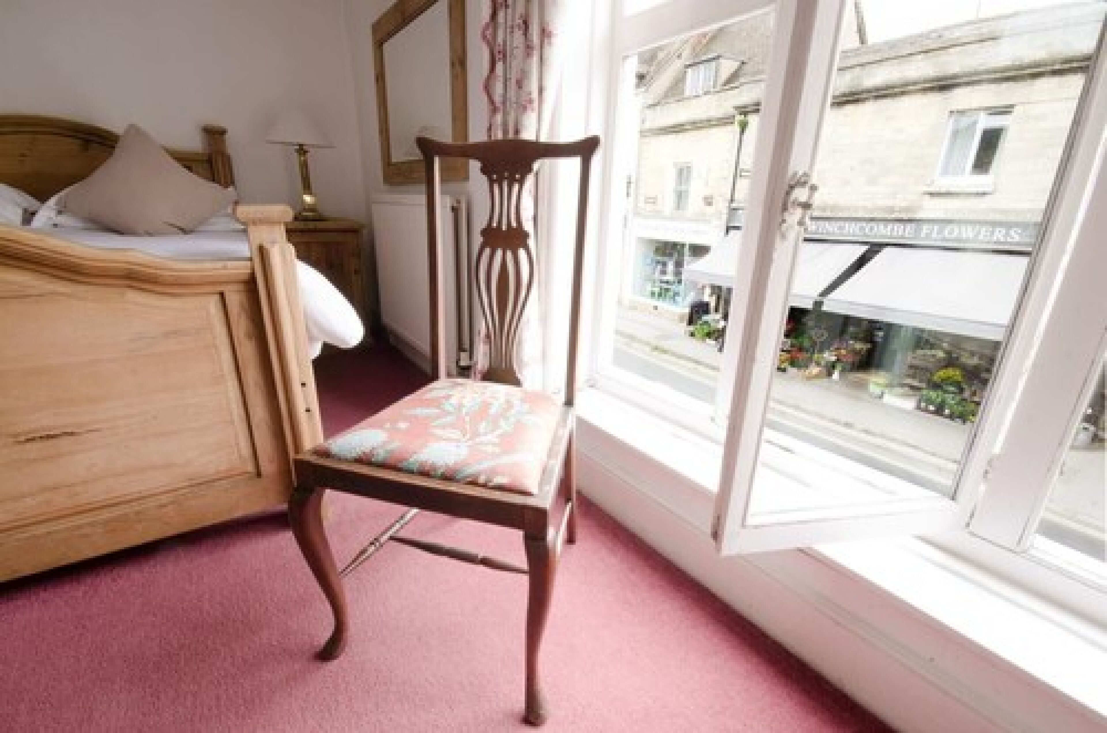 A bedroom with a wooden bed, a wooden chair with a cushion, a window with a view of a street, and a mirror on the wall.