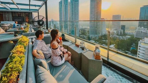 Een koppel zit op een balkon met uitzicht over de stad, een ventilator en een tafel met drankjes.