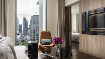 Quarto de hotel moderno com vista para a cidade, um sofá, uma poltrona, uma mesinha e uma TV fixada na parede.