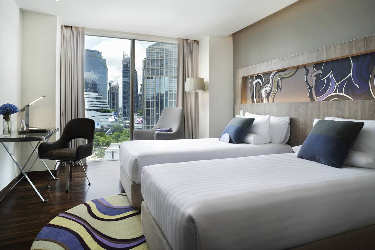 Habitació d'hotel amb dos llits, un escriptori amb una làmpada, una cadira, vista a la ciutat i una obra d'art abstracta moderna a la paret.