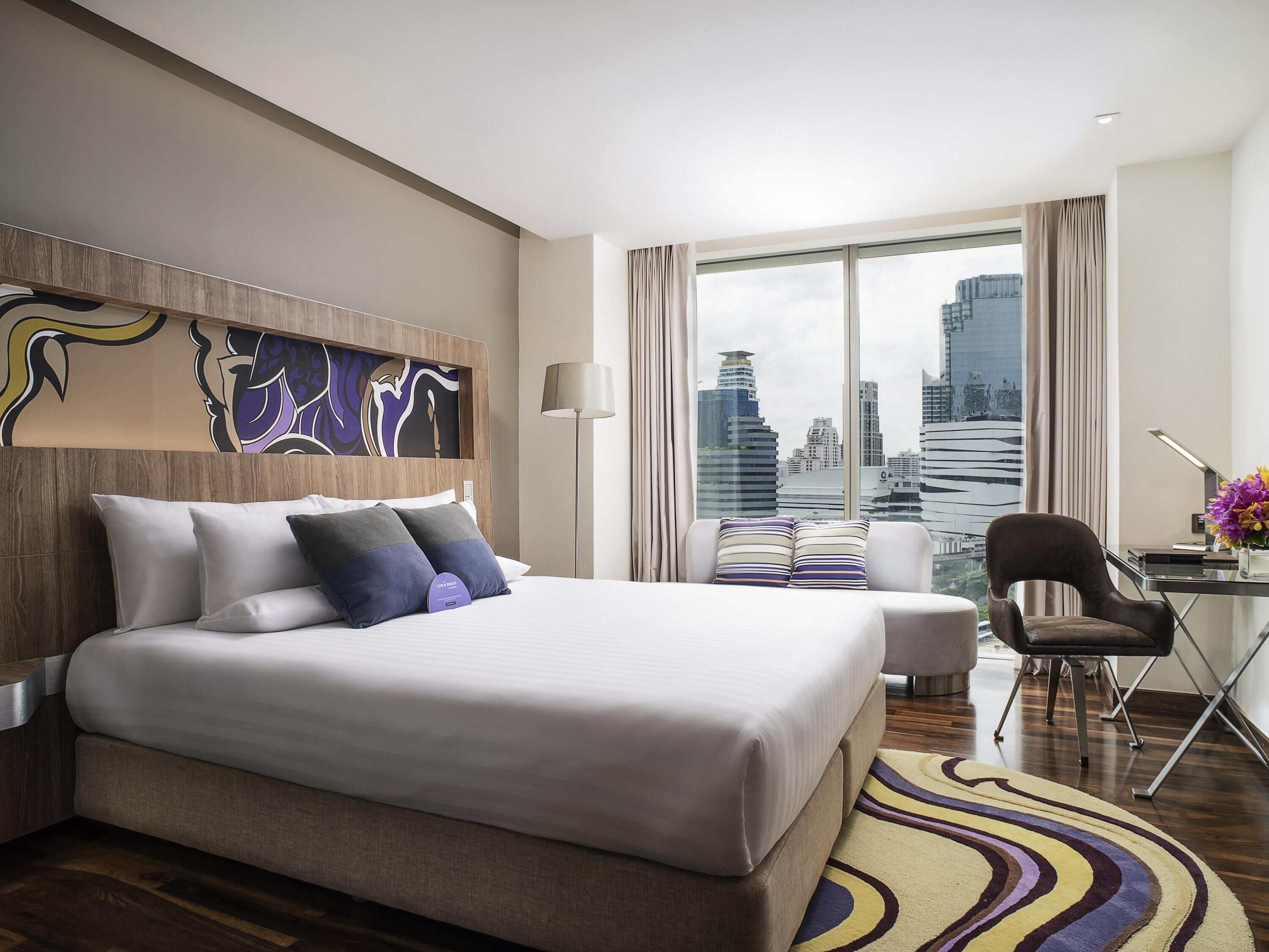 Una habitación de hotel moderna con una cama grande, un escritorio, una silla y vistas a la ciudad.