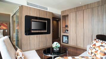 Habitación de hotel moderna con televisor de pantalla plana montado en la pared, una mesa de centro de vidrio con un jarrón de flores y un sofá con cojines estampados.