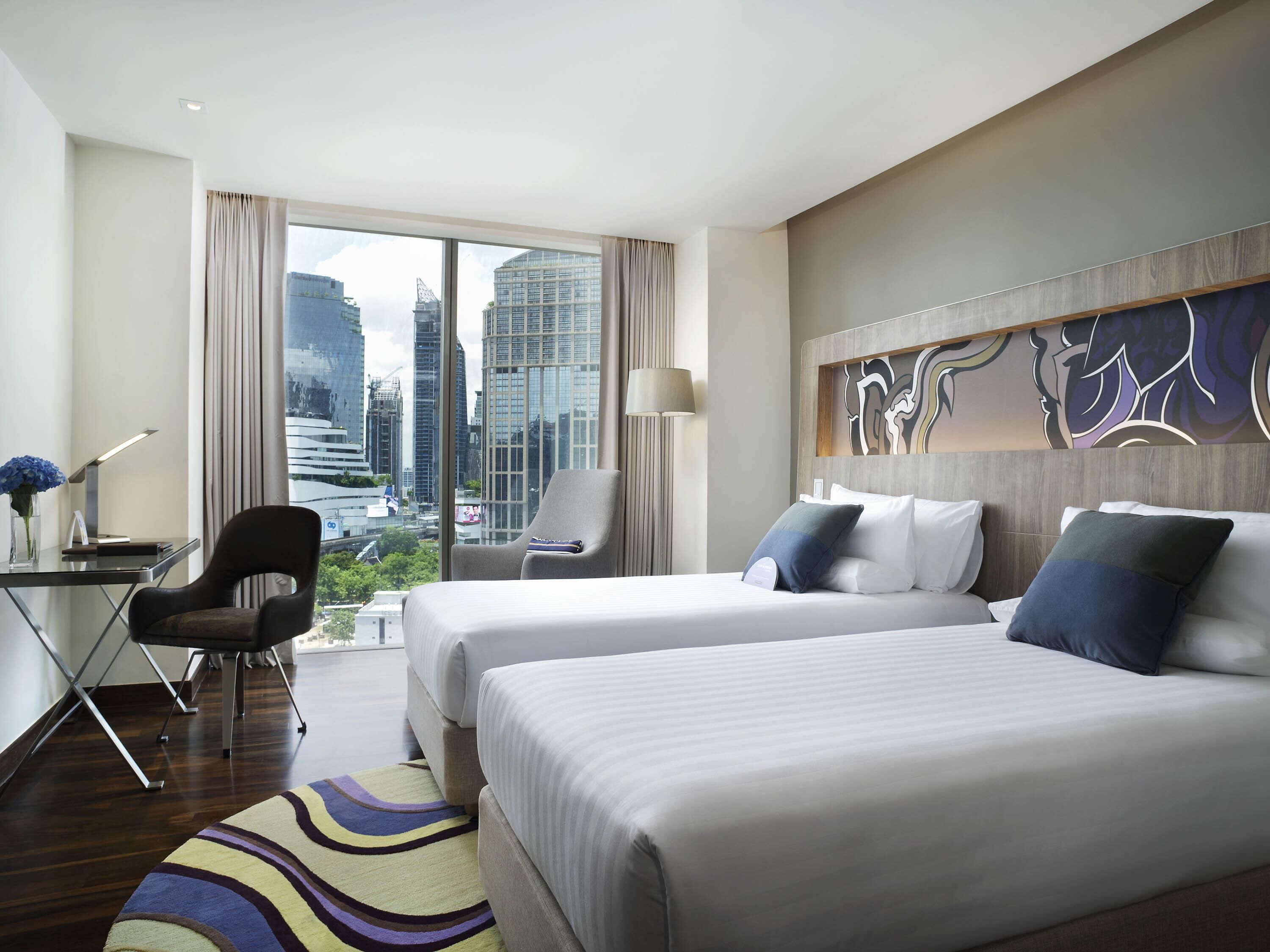 Habitación de hotel con dos camas, un escritorio, una silla y un ventanal con vistas a la ciudad.