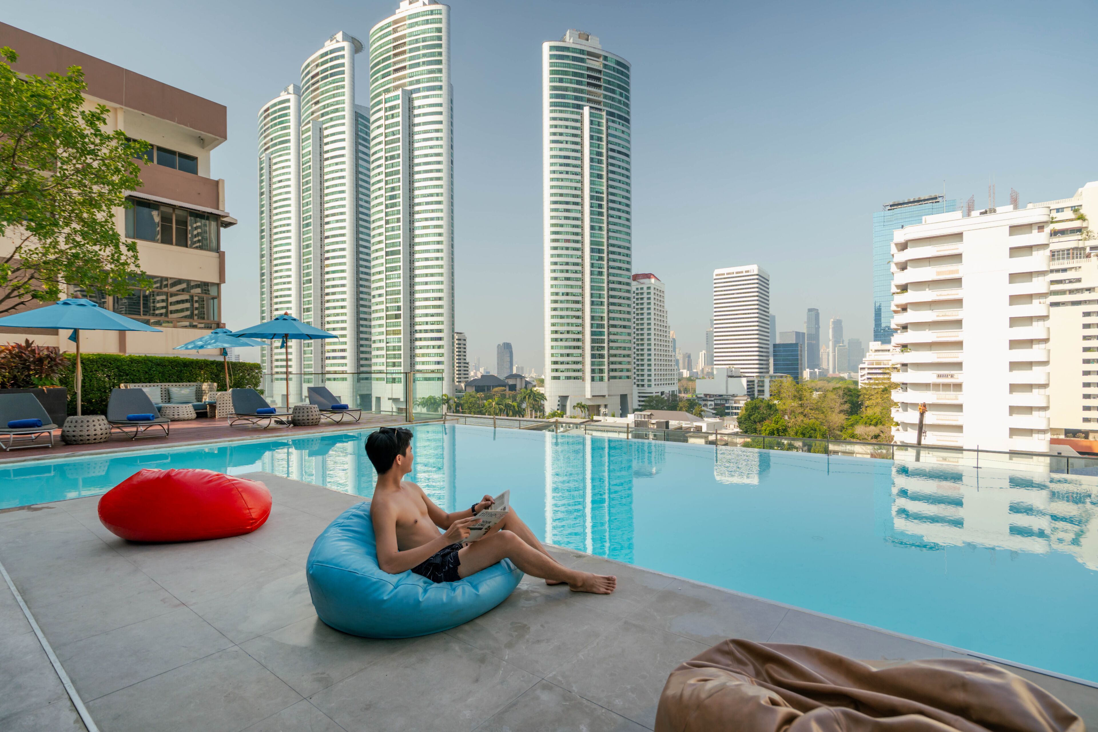 A poolside area with a person sitting on a bean bag, surrounded by modern buildings and a clear sky.