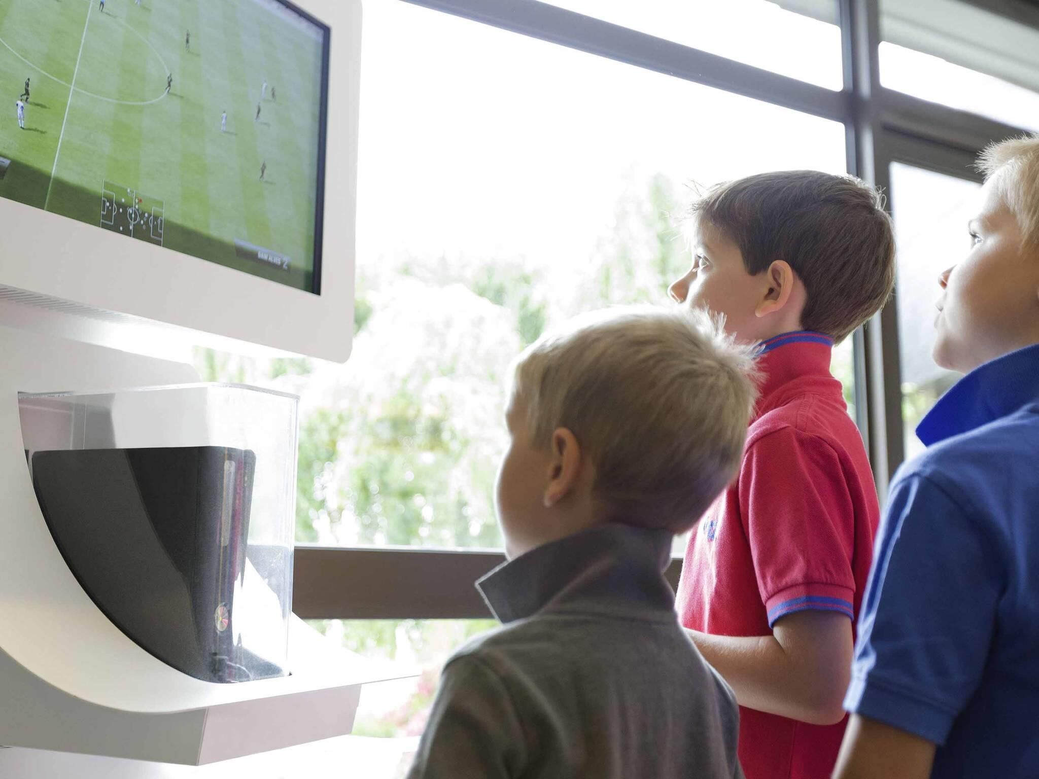 Un televisor montado en la pared mostrando un partido de fútbol, con tres niños mirando.