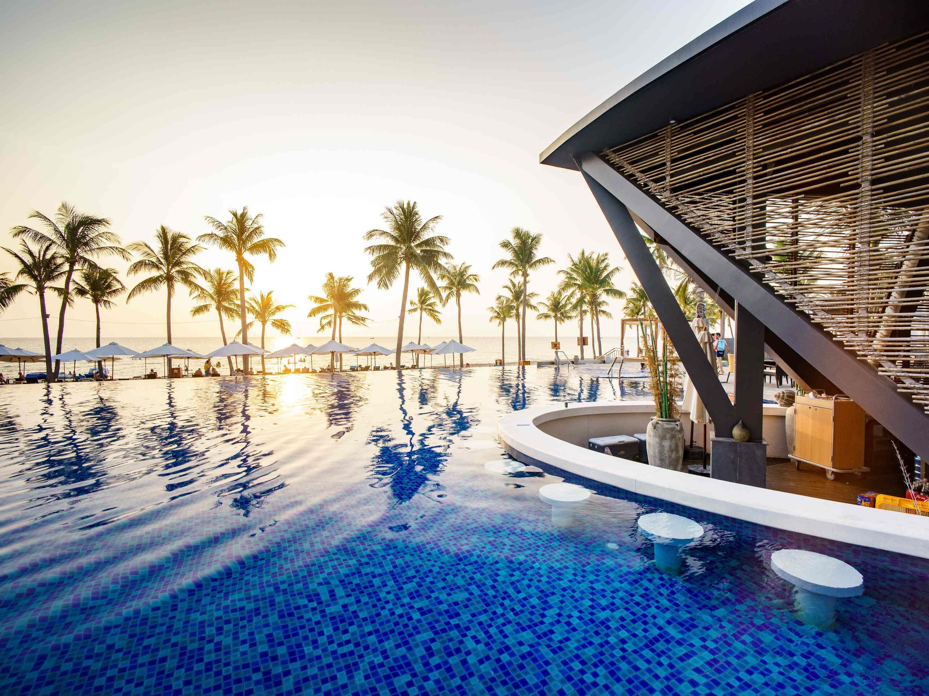 Piscina infinita con vista al océano y palmeras, una estructura de edificio moderna y un área de bar.