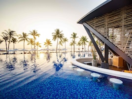 Piscina infinita con vista al océano y palmeras, una estructura de edificio moderna y un área de bar.