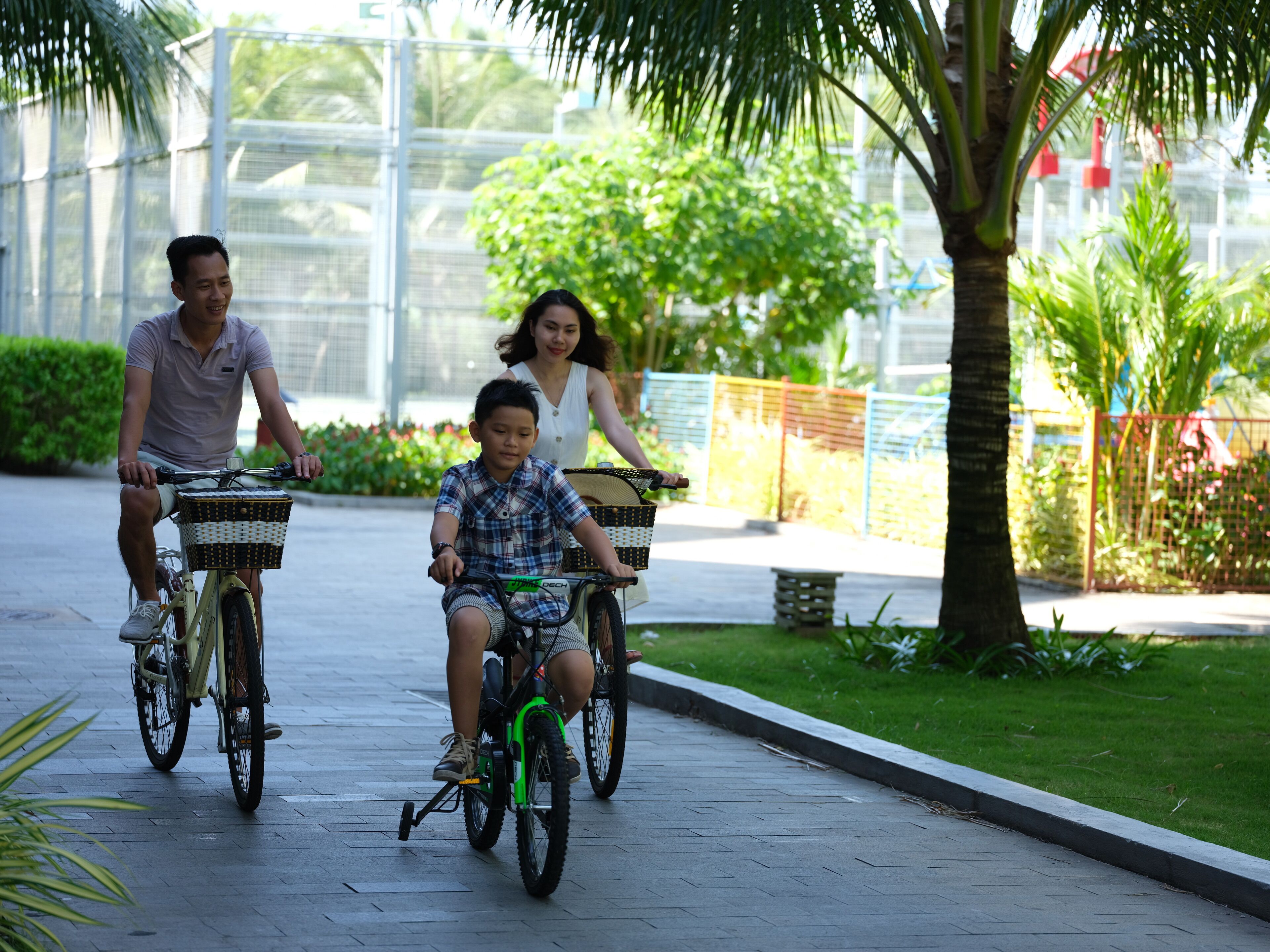 Three people riding bicycles on a paved path with greenery and a fence in the background.