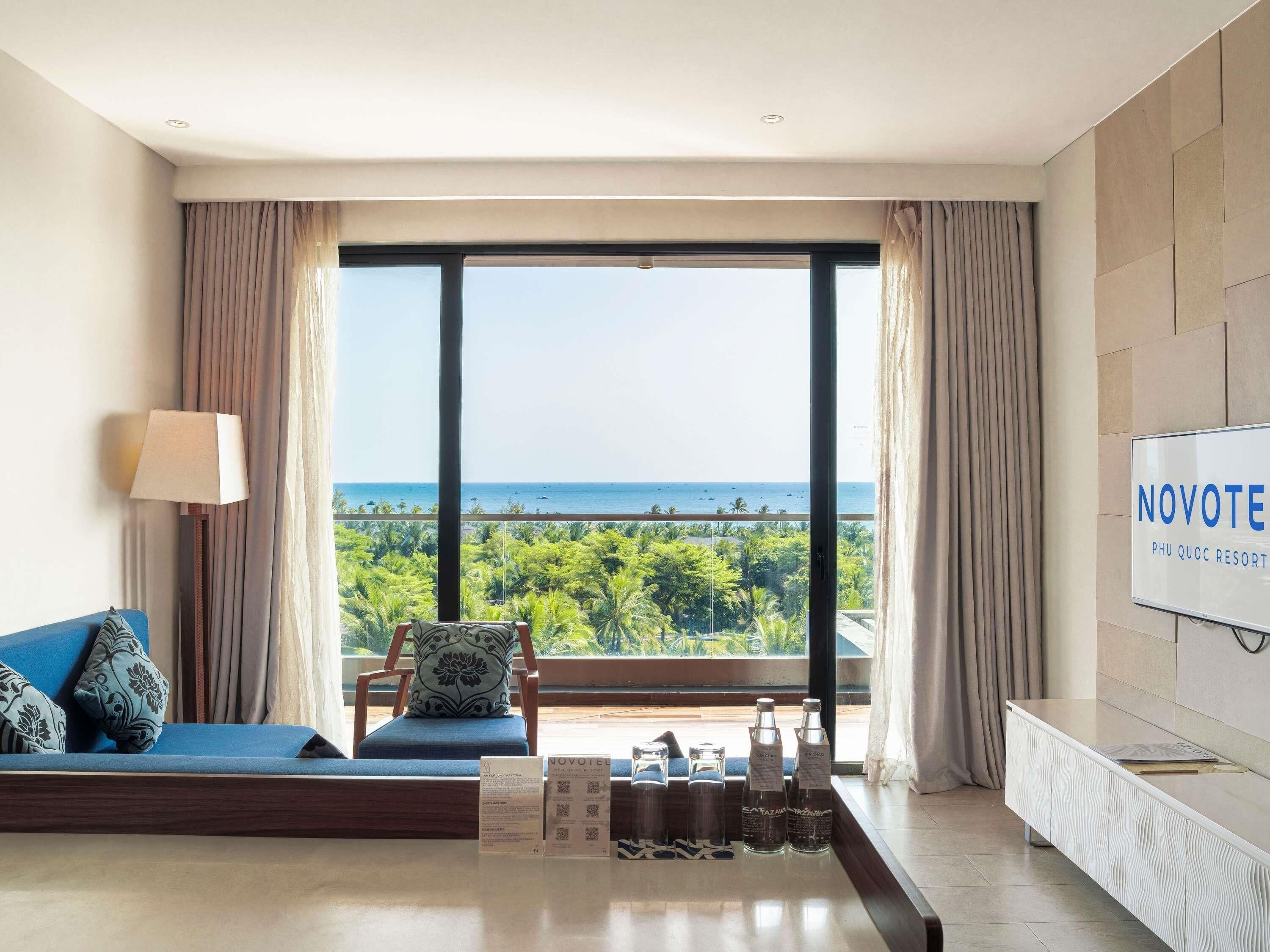 A modern hotel room with a large window, a sofa, a TV, and a minibar.