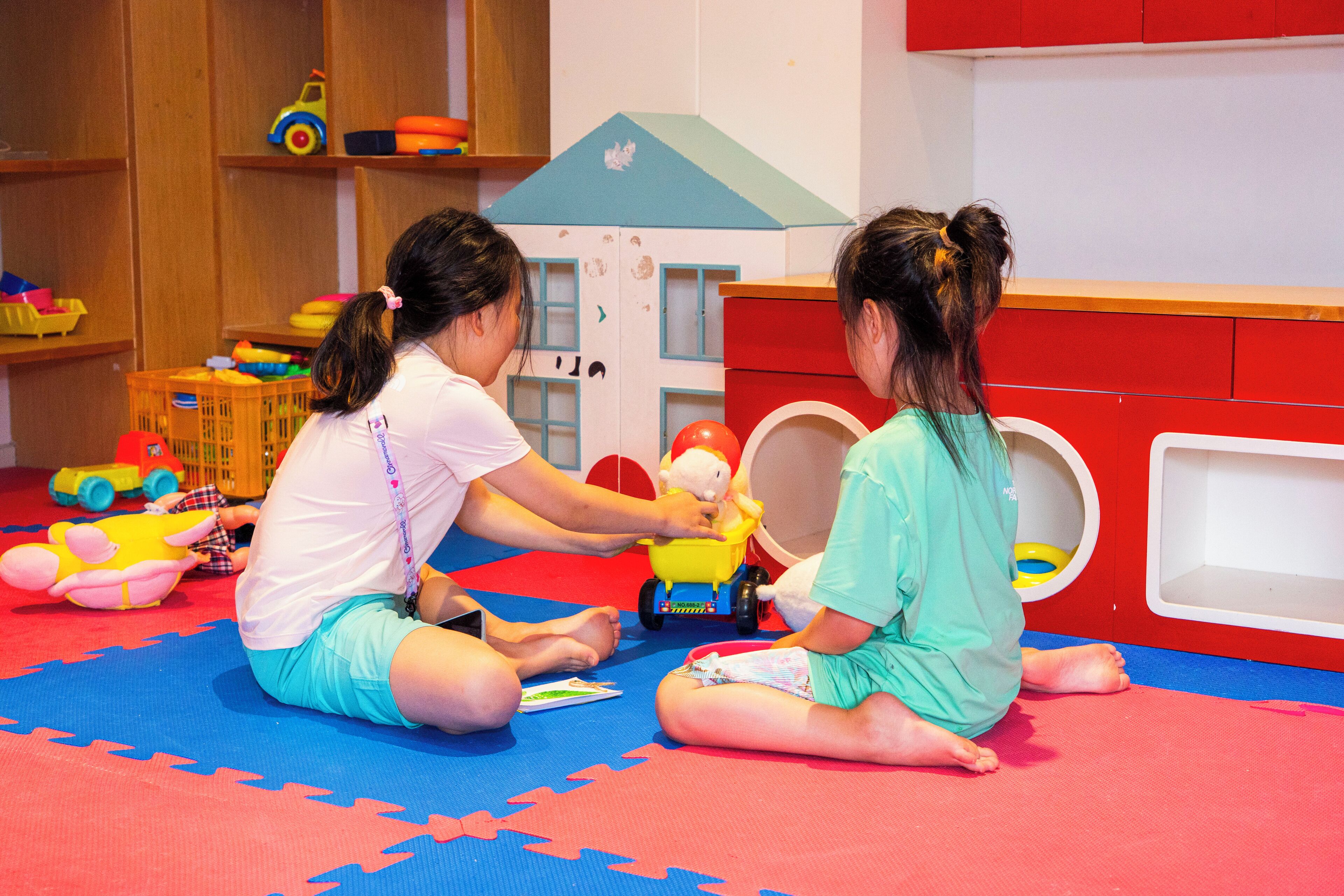 Two children playing with toys on a colorful mat in a room with wooden shelves and a red cabinet.
