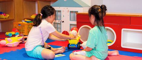 Two children playing with toys on a colorful mat in a room with wooden shelves and a red cabinet.